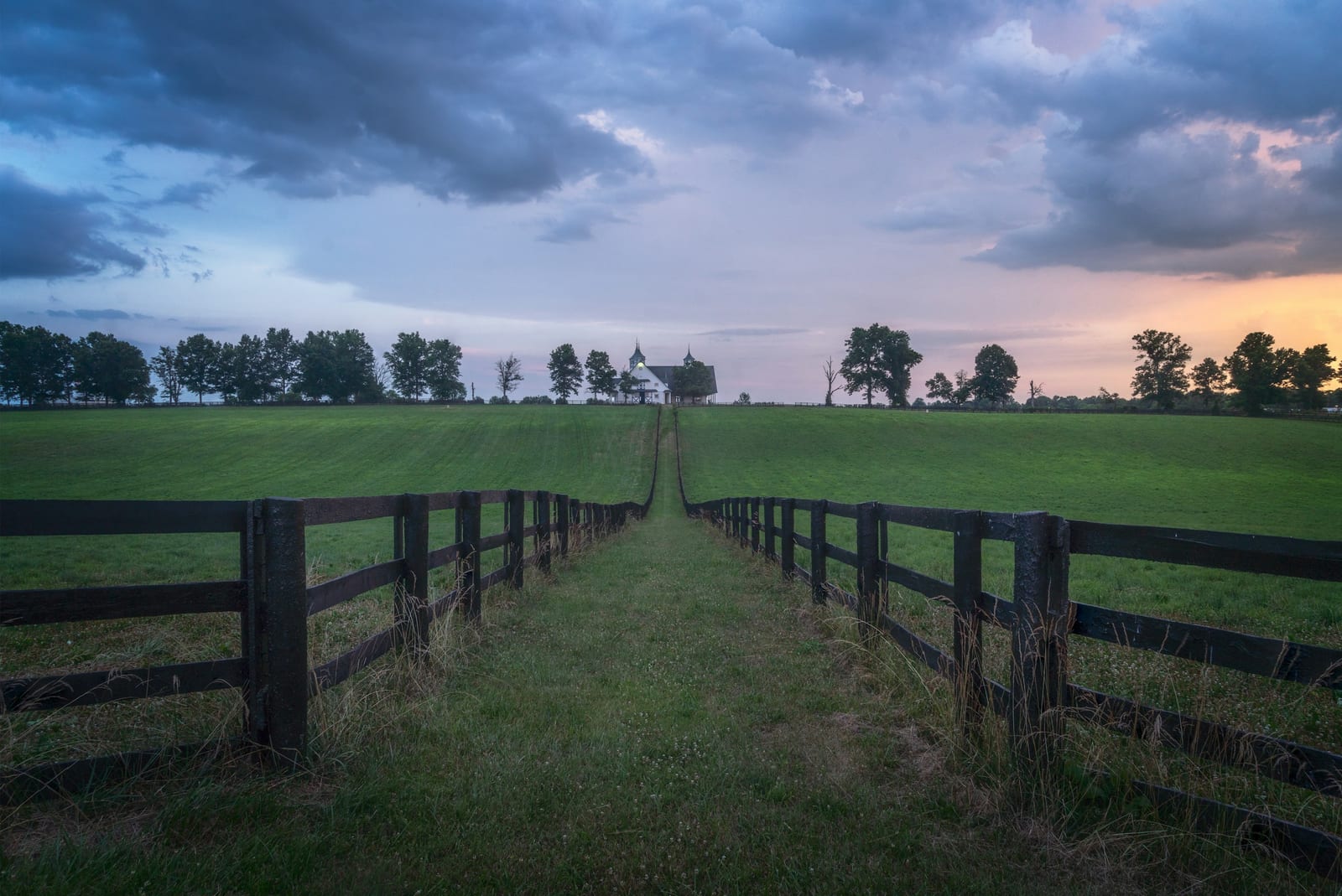 A grass lane between black plank fences leading uphill to a white horse barn under a dusk sky.
