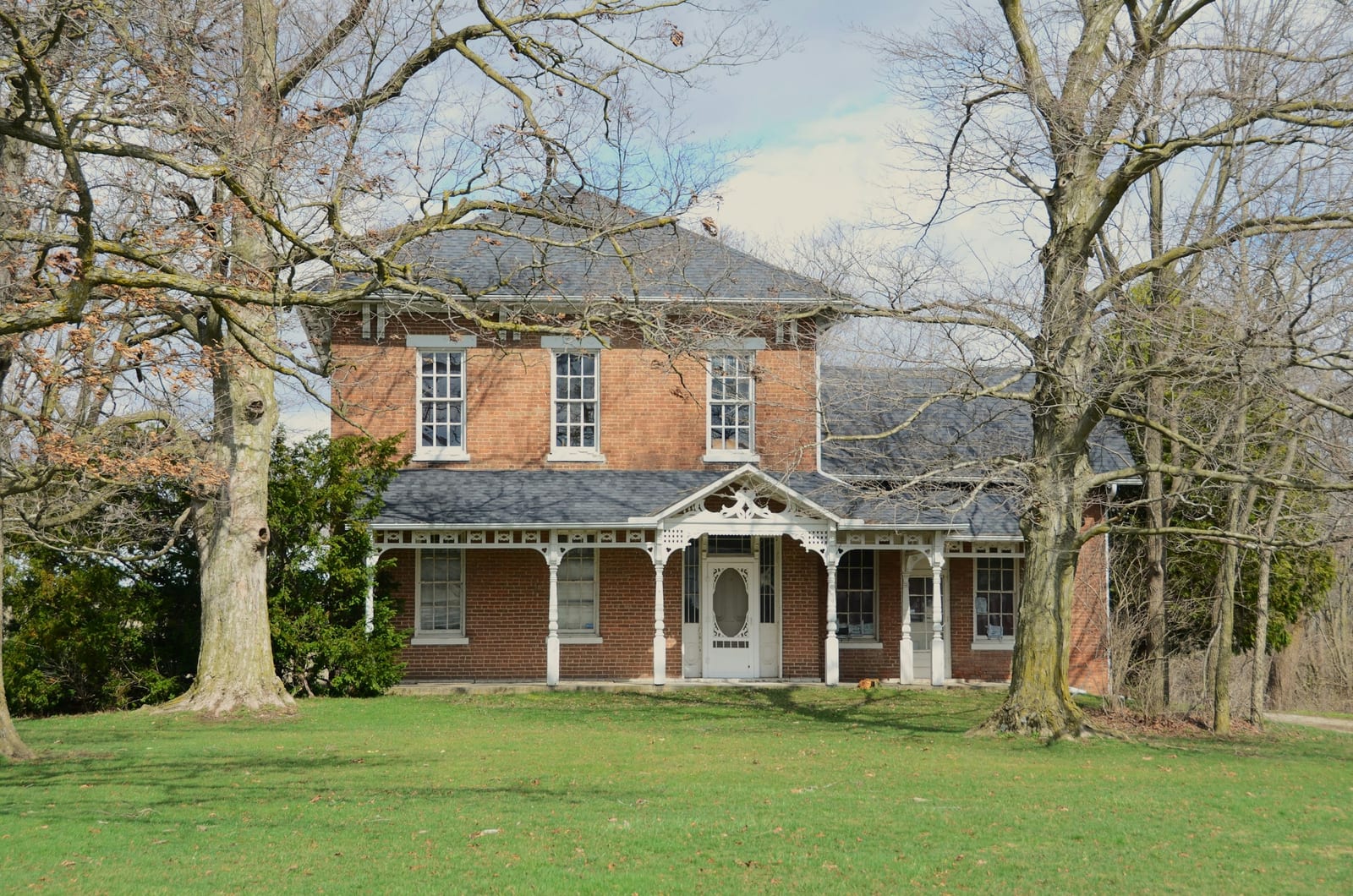 A two-story red-brick farmhouse with a white Victorian porch, framed by bare trees on a green lawn.
