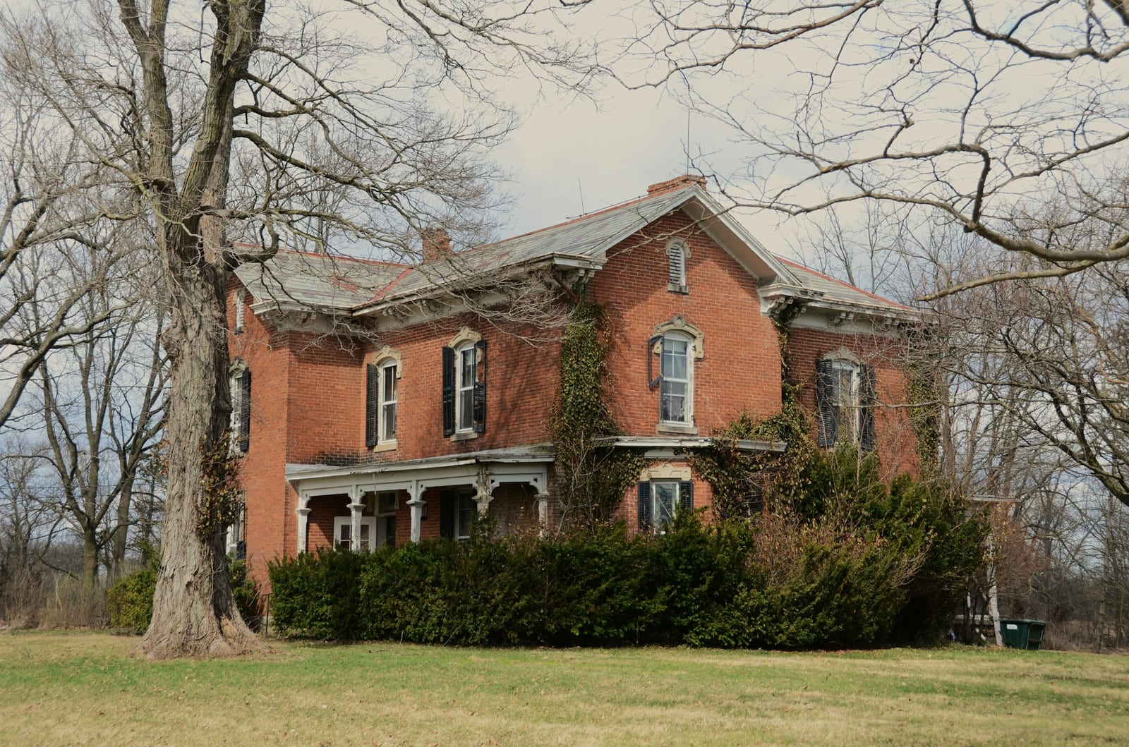 An older red-brick farmhouse with peeling trim and overgrown shrubs on a bare winter day.