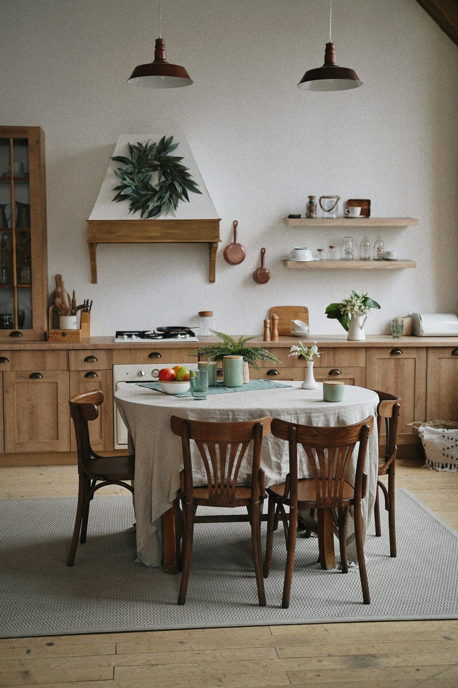 A warm farmhouse kitchen with wood cabinets, a round linen-covered table, and open shelving.